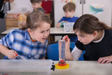 Auf dem Bild sind zwei Mädchen zu sehen, die begeistert an einem Experiment mit Wasser arbeiten. Sie untersuchen ein transparentes Behältnis mit Flüssigkeit, während andere Kinder im Hintergrund ebenfalls mit Experimentiermaterial beschäftigt sind.