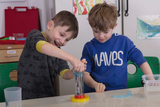Das Bild zeigt zwei Kinder, die begeistert ein Experiment mit einem transparenten Behälter durchführen, in dem Wasser und möglicherweise etwas anderes enthalten sind. Sie scheinen intensiv zu beobachten und miteinander zu interagieren, während sie das Experiment im Bildungsumfeld durchführen.