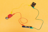 Das Bild zeigt einen einfachen elektrischen Schaltkreis mit einer Batterie, Drähten und einem kleinen Glockenmelder. Die bunten Kabel sind verbunden, um das Signal zur Glocke zu übertragen und verdeutlichen grundlegende Prinzipien der Elektrizität.