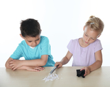 Auf dem Bild sind zwei Kinder zu sehen, die an einem Tisch arbeiten. Das Mädchen verwendet einen Stift, um mit einem schwarzen Stoffstück und kleinen Papierresten zu experimentieren, während der Junge aufmerksam zuschaut.