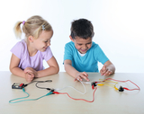 Das Bild zeigt zwei Kinder, die begeistert an einem Experiment mit elektrischen Bauteilen arbeiten. Sie verbinden Kabel und Komponenten, um eine Schaltung aufzubauen und das Prinzip elektrischer Stromverwendung spielerisch zu erlernen.