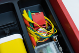 Das Bild zeigt eine Sammlung von Experimentiermaterialien in einem roten Aufbewahrungskasten. Zu sehen sind verschiedene Kabel mit Krokodilklemmen, eine kleine Batterie sowie weitere elektrische Komponenten, die für experimentelle Zwecke verwendet werden.