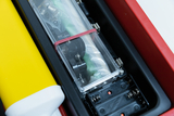 Das Bild zeigt einen geöffneten Experimentierkasten mit verschiedenen Komponenten, darunter ein transparentes Gehäuse mit einer Batterie und mechanischen Teilen. Daneben liegt ein gelbes Objekt, vermutlich ein Teil des Kits, das für Experimentierzwecke verwendet wird.