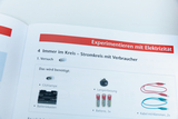 Das Bild zeigt eine Seite aus einem Lehrbuch über Experimente mit Elektrizität. Es listet Materialien auf, die für den Versuch eines Stromkreises mit Verbrauchern benötigt werden, wie Glühlampe, Lampenfassung und Batterien.