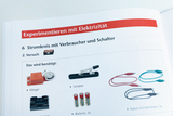 Das Bild zeigt eine Anleitung zum Experimentieren mit Elektrizität, speziell zum Aufbau eines Stromkreises mit Verbrauchern und Schaltern. Es listet die benötigten Materialien auf, darunter eine Klingel, Schalter, Batterien und Kabel.