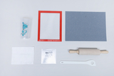 Das Bild zeigt eine Anordnung verschiedener Materialien für Experimente, darunter ein Becher mit einem lustigen Charakter, ein rotes Sieb, ein Stück blauer Pappe, ein Nudelholz, ein weißer Spatel und ein Informationsblatt. Diese Utensilien sind für schulische Projekte oder kreative Tätigkeiten geeignet.