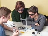 Das Bild zeigt drei Jugendliche, die konzentriert an einem Tisch mit Experimentiermaterialien arbeiten. Auf dem Tisch befinden sich eine Wärmequelle, Chemikalien in Behältern und eine Anleitung, während die Jugendlichen Sicherheitsbrillen tragen.