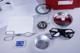 Das Bild zeigt eine Sammlung von Experimentierutensilien, darunter eine chemische Rohrzange, Schutzbrille, eine Spirituslampe und mehrere Behälter. Diese Ausrüstung wird häufig im naturwissenschaftlichen Unterricht verwendet.