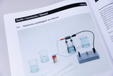 Das Bild zeigt ein Experiment zur elektrischen Leitfähigkeit von Wasser, das aus mehreren Bechergläsern und einem Gerät zur Messung besteht. Ein Batteriesatz ist mit Kabeln verbunden, um den Stromfluss zu demonstrieren.