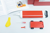 Das Bild zeigt Experimentiermaterialien, darunter eine rote Luftpumpe, einen gelben Ballon, eine kleine rote Kunststofffahrzeug sowie Zubehörteile. Im Hintergrund ist ein aufgeschlagenes Buch zu sehen, das Anleitungen für Experimente mit Wasser und Luft bietet.