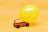 Das Bild zeigt ein gelbes Luftballon-Modell, das auf einem kleinen roten Wagen montiert ist. Der Wagen hat vier schwarze Räder und dient wahrscheinlich als Experimentiergerät, um physikalische Prinzipien wie Luftdruck und Bewegung zu veranschaulichen.