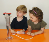 Auf dem Bild sind zwei Kinder zu sehen, die an einem Experimentiergerät arbeiten. Ein Kind hält eine Spritze, während das andere auf einen Zylinder zeigt, der mit Wasser gefüllt ist und ein kleines Fahrzeug oben hat.