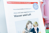 Das Bild zeigt ein Arbeitsheft mit dem Titel „Naturwissenschaften im Kindergarten“, das sich auf Experimente mit Wasser und Luft konzentriert. Es zeigt zwei Kinder, Anja und Leon, die gemeinsam an einem Experiment arbeiten.
