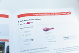 Das Bild zeigt eine Anleitung aus einem Experimentierbuch mit dem Titel „Luft kann Türen öffnen“. Es listet die benötigten Materialien wie einen Luftballon, eine Spritze und einen Schlauch auf, um das Experiment durchzuführen.