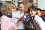 Das Bild zeigt eine Lehrerin, die mit zwei Kindern ein Experiment durchführt. Die Kinder sind begeistert und interagieren mit einem wissenschaftlichen Gerät, während ein weiteres Kind im Hintergrund aufmerksam zuschaut.