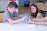 Auf dem Bild sind ein Junge und ein Mädchen zu sehen, die gemeinsam an einem blauen Ball arbeiten. Sie scheinen mit Eifer und Freude bei der Sache zu sein, während eine Hand eines Erwachsenen unterstützt.