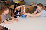 Das Bild zeigt zwei Kinder und eine Lehrerin, die gemeinsam an einem Experiment im Klassenraum arbeiten. Das Mädchen bedient eine Apparatur, während die Lehrerin unterstützend eingreift, und der Junge beobachtet die Aktivität aufmerksam.