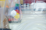 Das Bild zeigt eine transparente Box mit verschiedenen Experimentiermaterialien, darunter Becher, Batterien und farbige Bauklötze. Zusätzlich sind Schlauchstücke und weitere Vorrichtungen für wissenschaftliche Experimente enthalten.