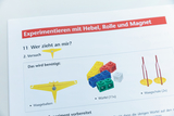Das Bild zeigt eine Anleitung für ein Experiment, das sich mit Hebel, Rollen und Magneten beschäftigt. Es listet die benötigten Materialien auf, darunter einen Waagenbalken, diverse farbige Würfel und zwei Waagschalen.