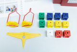 Das Bild zeigt verschiedene experimentelle Lernspielzeuge, darunter bunte Würfel in unterschiedlichen Farben und Formen sowie zwei rote Halterungen mit gelben Elementen. Im Hintergrund sind Arbeitsmaterialien und Anleitungen für das Experimentieren im Bildungsbereich sichtbar.