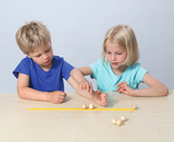 Auf dem Bild sind zwei Kinder zu sehen, die an einem Tisch sitzen und sich mit Holzperlen und einem gelben Lineal beschäftigen. Sie scheinen gemeinsam ein Experiment durchzuführen, wobei eines der Kinder mit den Perlen spielt, während das andere genau hinschaut.