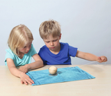 Das Bild zeigt zwei Kinder, die konzentriert mit einem Experimentiergerät spielen. Auf einem blauen Tuch liegt eine Kugel in einem behälter, während eines der Kinder an einem Faden zieht, um eine physikalische Experimente durchzuführen.