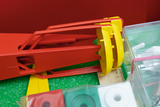 Das Bild zeigt verschiedene Experimentiermaterialien für den Bildungsbereich, darunter ein rot-gelbes Gerät und eine Auswahl an farbigen Platten. Diese Materialien sind auf einer grünen Unterlage angeordnet und scheinen für praxisnahe Lernaktivitäten konzipiert zu sein.