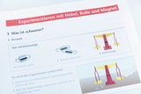 Das Bild zeigt eine Seite aus einem Experimentierhandbuch, die sich mit dem Thema Hebel und Gewichte beschäftigt. Es enthält Anleitungen für ein Experiment zur Bestimmung, welches Gewicht schwerer ist, und präsentiert die benötigten Materialien sowie eine Skizze der Balkenwaage.