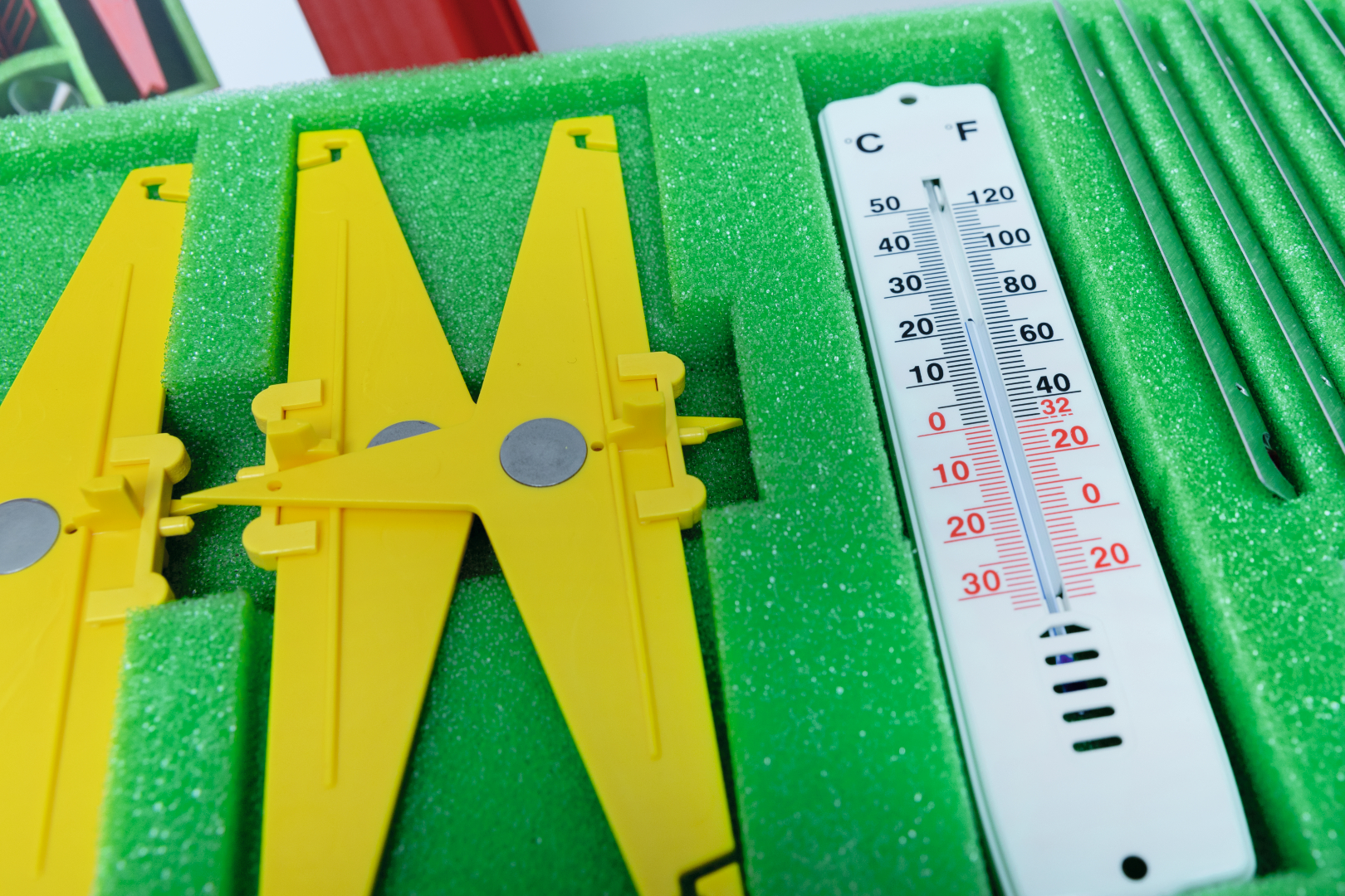 Das Bild zeigt eine sorgfältig angeordnete Sammlung von Experimentierwerkzeugen, darunter gelbe Schneider und ein Thermometer, die auf einer grünen Schaumstoffunterlage liegen. Diese Materialien sind für den Einsatz im Bildungswesen konzipiert und fördern praktische Lernexperimente.