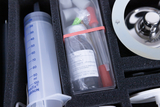 Das Bild zeigt eine Box mit verschiedenen Experimentiermaterialien, darunter eine Spritze und Reagenzgläser. Die Aufbewahrung erfolgt in einem schaumstoffgepolsterten Innenraum, der einen sicheren Transport der Utensilien ermöglicht.