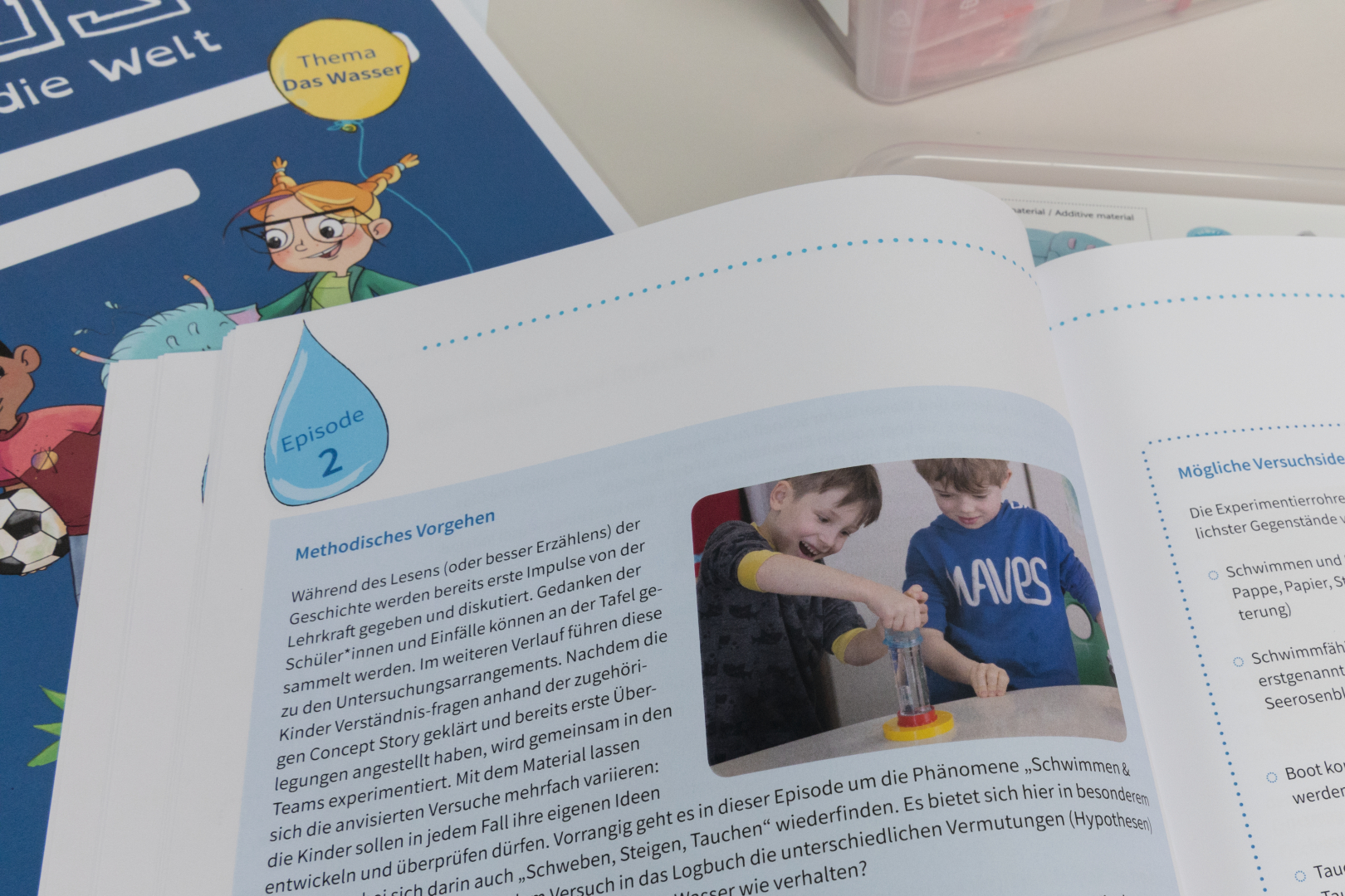 Das Bild zeigt eine Lehrbuchseite mit dem Thema "Das Wasser", auf der Kinder an einem Experiment teilnehmen, das sich mit den Phänomenen Schwimmen und Tauchen beschäftigt. In den danebenstehenden Notizen werden mögliche Versuchsaufbauten und methodische Vorgehensweisen erläutert.