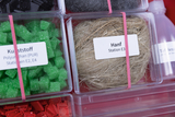 Das Bild zeigt verschiedene Materialien in transparenten Behältern, darunter grüne Kunststoffstücke und Hanf. Jedes Behältnis ist mit einem Etikett versehen, das die Inhaltsstoffe und die zugehörigen Stationen im Experimentierbedarf angibt.