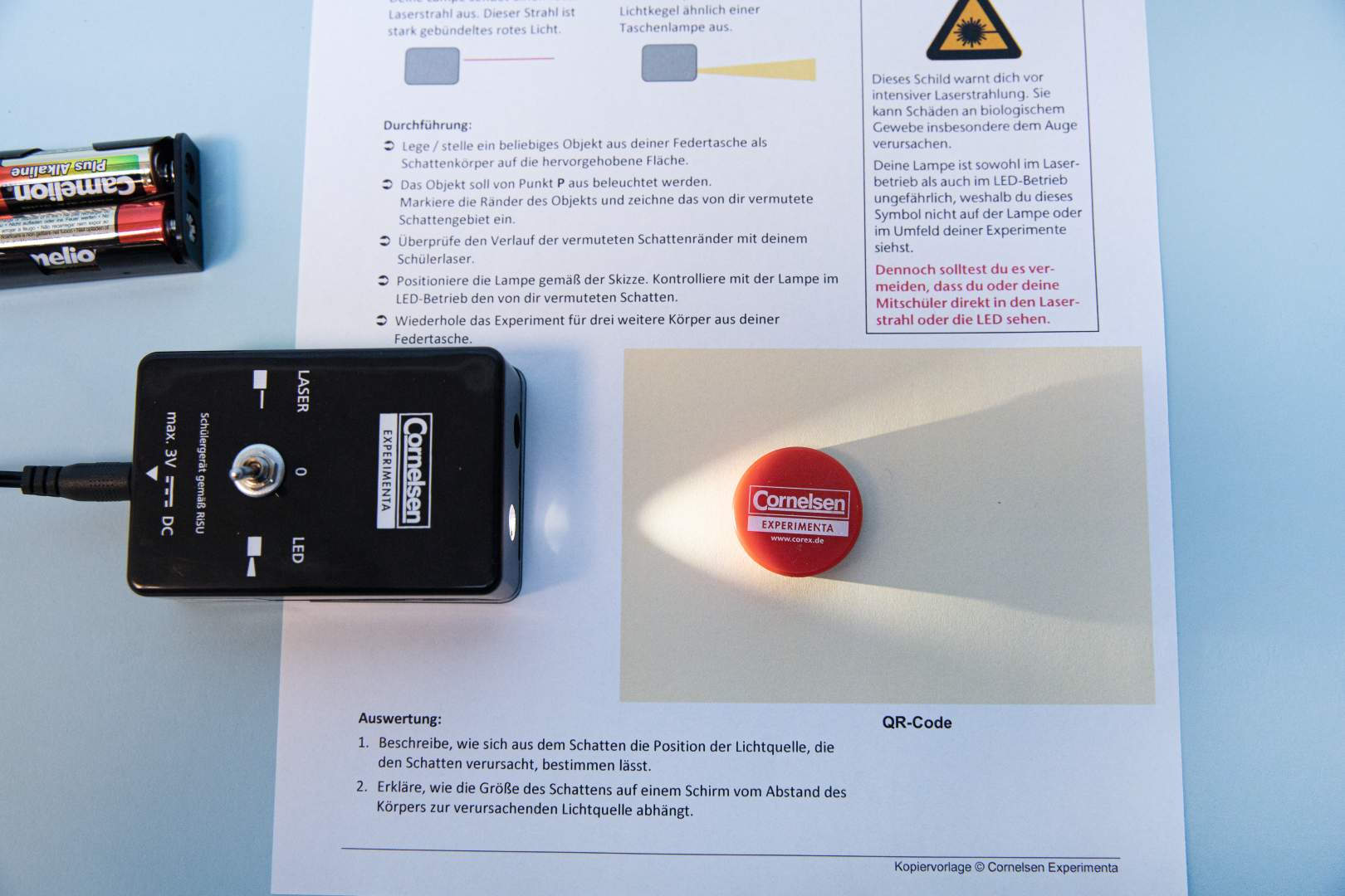 Das Bild zeigt ein Experimentierset mit einem Lasergerät und einer Anleitung, die die Durchführung eines Experiments zur Schattenbildung beschreibt. Auf einem weißen Blatt ist ein rotes Spielzeug oder eine Marke abgebildet, auf die das Licht des Lasers projiziert wird.