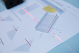 Das Bild zeigt einen transparenten Prisma, der auf einem Arbeitsblatt liegt, welches Diagramme zur Brechung von Lichtstrahlen enthält. Rote Laserlinien verdeutlichen den Verlauf des Lichtes durch das Prisma und die entsprechenden Winkel an den Punkten A, B und C.