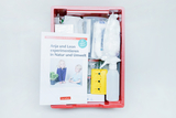 Das Bild zeigt eine rote Box mit Experimentiermaterialien für den Kindergarten, die verschiedene Utensilien wie Flaschen, Sprühgeräte und verschlossene Behälter enthält. Auf der Oberseite liegt ein Buch mit dem Titel „Anja und Leon experimentieren in Natur und Umwelt“, das Anleitungen bietet.