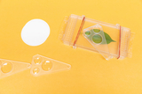 Das Bild zeigt verschiedene experimentelle Geräte auf einem gelben Untergrund, darunter einen Behälter mit einem grünen Blatt und zwei vergrößerten Linsen. Außerdem liegen zwei transparente Möbelstücke und eine weiße runde Karte daneben.