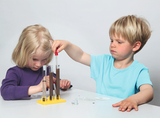 Das Bild zeigt zwei Kinder, die mit Reagenzgläsern auf einem Tisch experimentieren. Ein Mädchen mit blonden, kurzen Haaren konzentriert sich auf die Gläser, während ein Junge mit blonden Haaren ein Pipettiergerät benutzt, um Flüssigkeit in ein Glas zu geben.