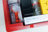 Das Bild zeigt einen roten Behälter mit verschiedenen experimentellen Materialien, darunter Behälter für kleine Teile, rote Kugeln und verschiedene Werkzeugteile. Auf dem Behälter sind auch Sicherheits- und Herstellungsinformationen sichtbar.