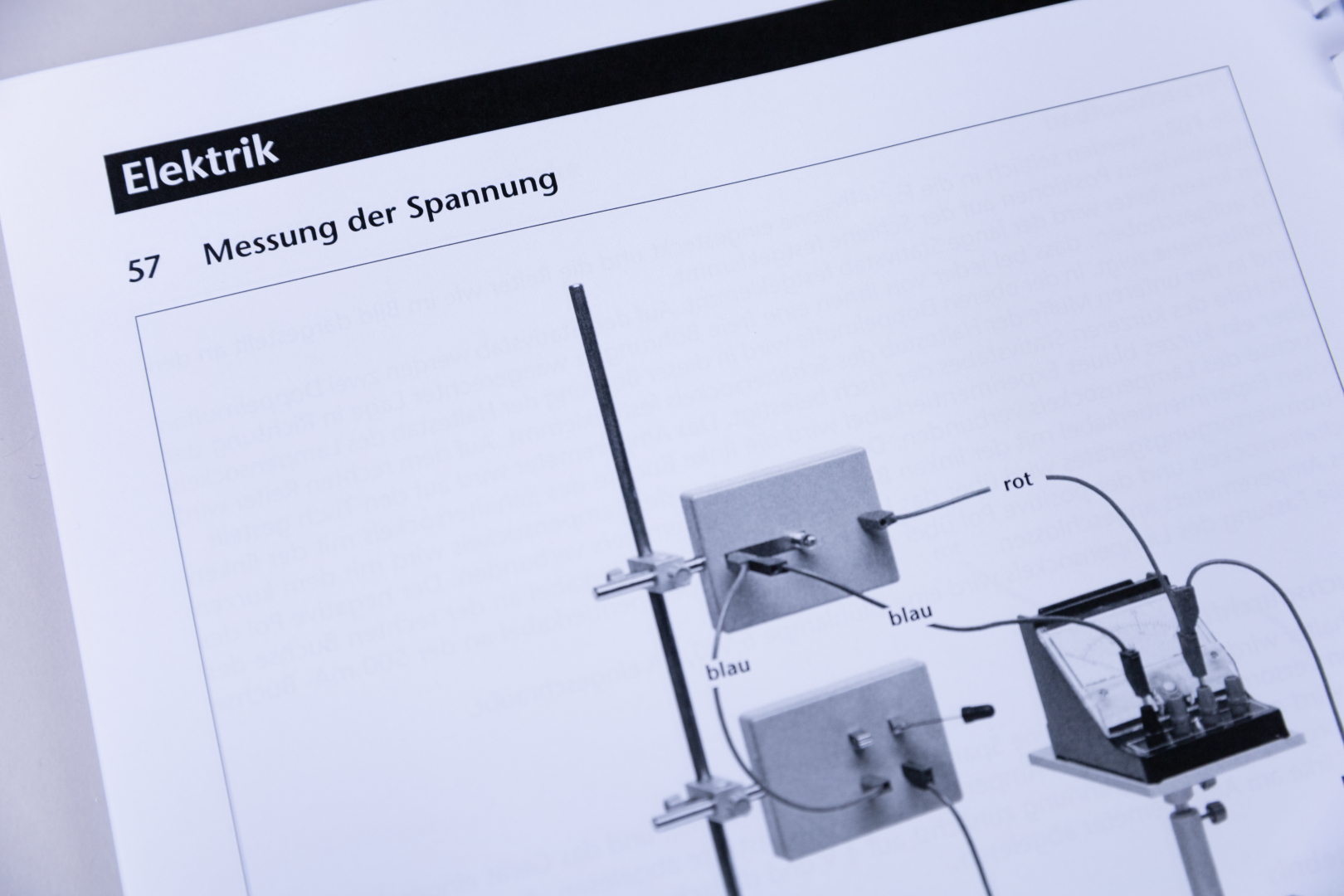 Das Bild zeigt eine Abbildung aus einem Lehrbuch über Elektrik, die die Messung von Spannung illustriert. Zwei experimentelle Geräte sind verbunden, wobei Kabel in den Farben rot und blau verwendet werden, um die Spannung zu messen.