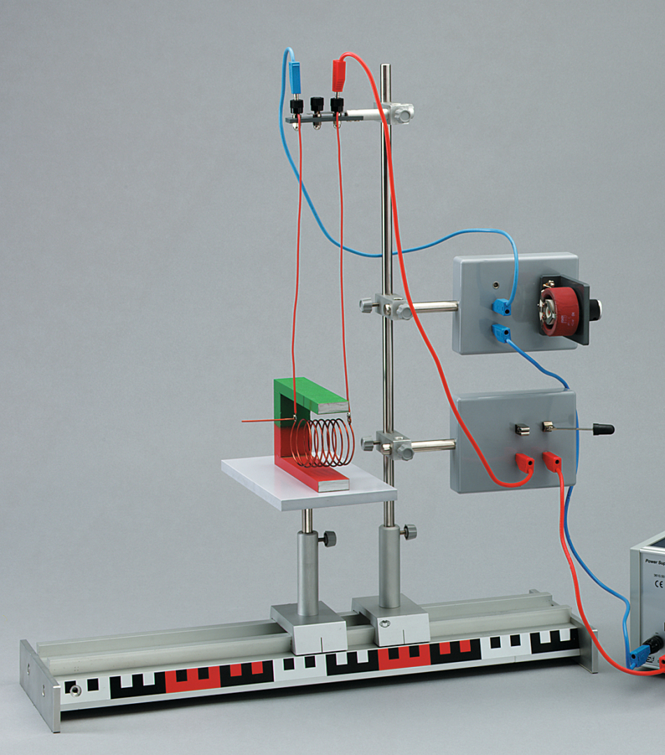 Das Bild zeigt ein Experimentiergerät zur Demonstration elektromagnetischer Prinzipien. Es besteht aus einer Anordnung mit Spulen, einem Magneten und verschiedenen Anschlüssen für elektrische Leitungen, die auf einer stabilen Plattform montiert sind.