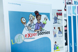 Das Bild zeigt ein Plakat mit dem Titel "Luft eXperTeenies", das zwei comicartige Figuren darstellt: einen Jungen mit einer Uhr und ein Mädchen mit einem Erlenmeyerkolben. Es ist Teil einer Handreichung für Experimentierbedarf, wahrscheinlich im schulischen Kontext.