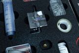 Das Bild zeigt eine Sammlung von Experimentiermaterialien, die in einem Schaumstoff-Organizer angeordnet sind. Zu sehen sind unter anderem eine Spritze, verschiedene Teile, ein Glasbehälter mit einer chemischen Substanz und kurze Kabel.