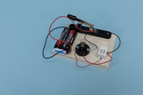 Das Bild zeigt eine einfache Schaltung bestehend aus zwei Batterien, einer Lampe und einem Schalter, die auf einem Holzbrett montiert sind. Diese Konstruktion dient vermutlich zur Durchführung von Experimenten im Bildungsbereich, um Grundlagen der Elektrizität zu vermitteln.