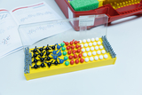 Das Bild zeigt ein Molekülbaukasten-Set, das zur Visualisierung von Molekülstrukturen verwendet wird. Bunte Elemente sind auf einer gelben Platte angeordnet, während im Hintergrund ein Handbuch mit chemischen Strukturen zu sehen ist.