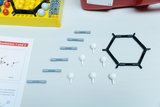 Das Bild zeigt ein Set mit Modellbaukästen zur Demonstration von Molekülstrukturen, bestehend aus verschiedenen farbigen Kugeln und Verbindungsstücken in einer gelben Box sowie einer Anleitung. Neben dem Set sind einige Molekülmodelle, darunter ein Hexagon, auf dem Tisch arrangiert.