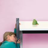 Das Bild zeigt ein Kind, das seitlich an einem Tisch lehnt und aufmerksam auf ein grünes Plüschtier schaut, das auf der Tischplatte sitzt. Der Hintergrund ist in einem sanften Rosa gehalten, was eine freundliche und einladende Atmosphäre schafft.