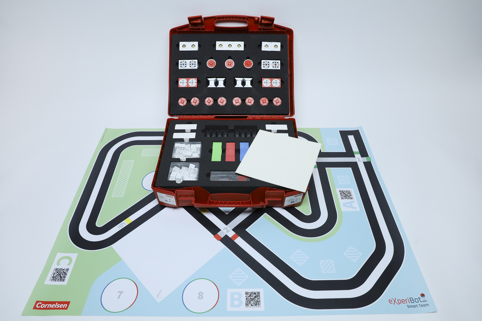 Offener Experimentierkoffer mit dem dazugehörigen Spielplan, auf dem die Fabrik aufgebaut werden kann. Der Spielplan ist ausgebreitet und der offene Koffer steht auf dem Spielplan. 