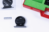 Das Bild zeigt ein schwarzes Experimentiergerät, das auf einem weißen Hintergrund liegt, neben einer rot-grünen Box, die mit Schaumstoff ausgekleidet ist. Ein Blatt Papier mit dem Logo "Cornelsen" und einer Illustration des Geräts liegt ebenfalls daneben.