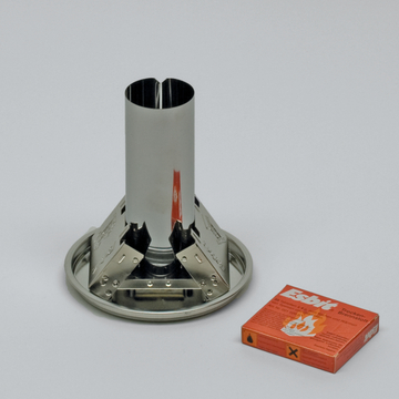 Das Bild zeigt einen Metall-Brenner mit einem hohlen, zylindrischen Körper und einer stabilen Basis, die für Experimente verwendet wird. Daneben liegt eine Packung Esbit-Trockenbrennstoff, der zur Erzeugung von Wärme während des Experimentierens dient.