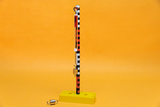 Das Bild zeigt eine experimentelle Vorrichtung mit einem senkrechten Stab, der eine Federwaage und einen schweren Block hält. Die Basis ist in leuchtendem Gelb und bietet Stabilität für das Experiment, das wahrscheinlich physikalische Prinzipien veranschaulicht.